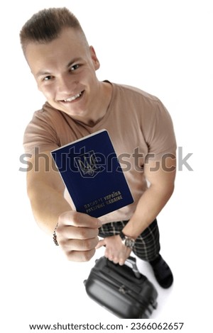 Above view of a handsome young man in a beige T-shirt and plaid pants posing with a suitcase and showing a Ukrainian passport isolated on white background