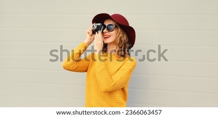 Autumn portrait of beautiful young woman photographer with film camera taking picture wearing hat, yellow sweater on gray background