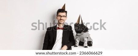 Image of handsome young man celebrating birthday with cute black pug in party costume and cone on head, standing over white background.