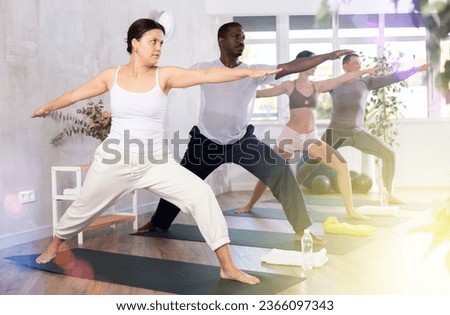 Positive young asian woman practicing Virabhadrasana known as Warrior Pose during group yoga training in fitness studio..