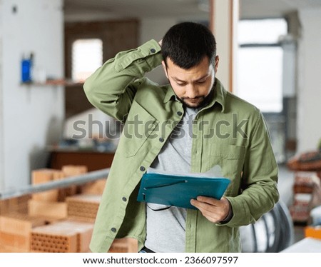 Puzzled and confused young bearded man standing in his house with papers in hands during renovations, determining scope of work Royalty-Free Stock Photo #2366097597