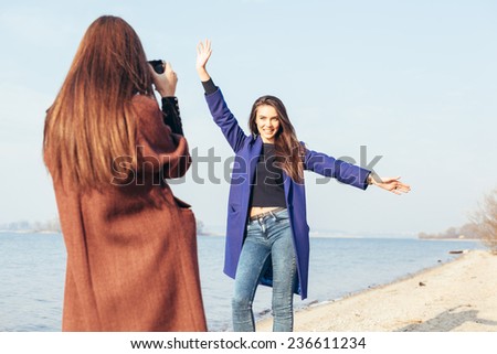 Beautiful young girl taking pictures her cheerful girlfriend on the beach in front of the sea. Outdoor lifestyle portrait