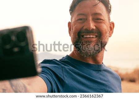 Happy man outdoor taking selfie picture using smartphone mobile and smiling at the camera. Online sharing social life. People and travel pictures. White sky background scenic nature place country side