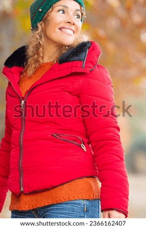 Young happy cheerful adult woman posing outdoor and enjoying leisure activity wearing a red warm jacket. Autumn colors and trees in background. Cute middle age female people at the park smiling