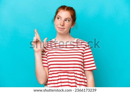 Young reddish woman isolated on blue background with fingers crossing and wishing the best