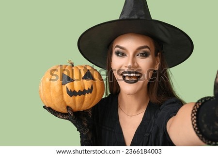 Young woman dressed for Halloween as witch with pumpkin taking selfie on green background, closeup