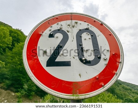 Round traffic road sign. Speed limit 20 km. Black color, number twenty. Red circle. Bullet holes. Nature, and washed white sky.