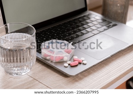 Organizer weekly shots with glass of water on workplace background and laptop Closeup of medical pill box with doses of tablets for daily take medicine with white pink drugs and capsules. Health care