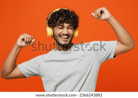 Young smiling fun cool happy Indian man he wears t-shirt casual clothes listen to music in headphones raise up hands dance isolated on orange red color background studio portrait. Lifestyle concept