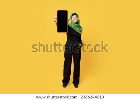 Full body happy young arabian asian muslim woman wears green hijab abaya black clothe hold use blank screen mobile cell phone isolated on plain yellow background. People uae islam religious concept