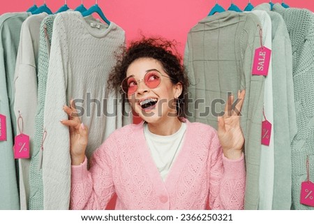 Young smiling minded dreamful happy female costumer woman wear sweater stand near clothes rack with tag sale in store showroom take off glasses look overhead isolated on plain pink background studio.