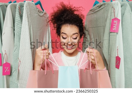 Young smiling costumer woman 20s in sweater stand near clothes rack with tag sale in store showroom hold look at package bags with purchases after shopping isolated on plain pink background studio.