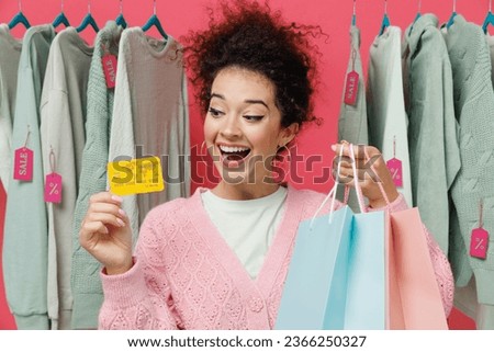 Young surprised costumer woman in sweater stand near clothes rack with tag sale in store showroom hold package bags with purchases after shopping credit card isolated on plain pink background studio