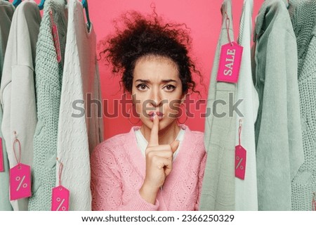 Young secret costumer woman 20s in sweater stand near clothes rack with tag sale in store showroom say hush be quiet with finger on lips shhh gesture isolated on plain pink background studio portrait