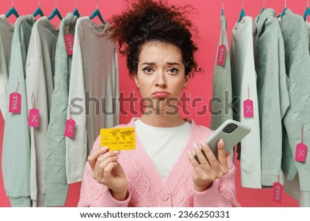 Young sad displeased woman in sweater stand near clothes rack with tag sale in store showroom using mobile cell phone hold credit bank card shopping online isolated on plain pink background studio.