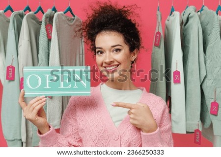 Young smiling female costumer woman 20s in sweater stand near clothes rack with tag sale in store showroom hold point finger on card sign with open title text isolated on plain pink background studio