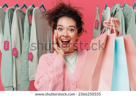 Young costumer woman 20s in sweater stand near clothes rack with tag sale in store showroom show package bags with purchases after shopping hold face isolated on plain pink background studio portrait