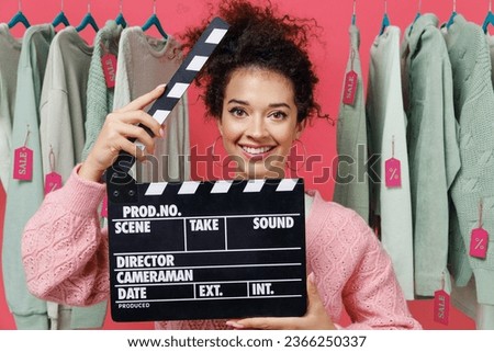 Young female costumer woman 20s wear sweater stand near clothes rack with tag sale in store showroom holding classic black film making clapperboard isolated on plain pink background studio portrait