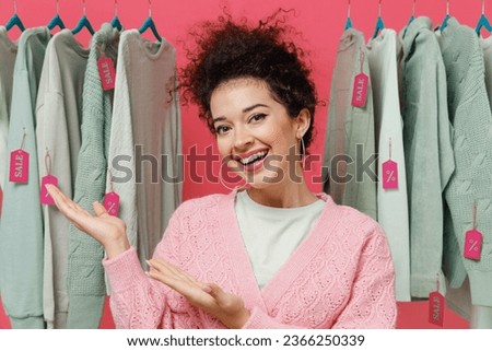 Young smiling happy cheerful female costumer woman 20s wear sweater stand near clothes rack with tag sale in store showroom point hands on jumpers isolated on plain pink background studio portrait.