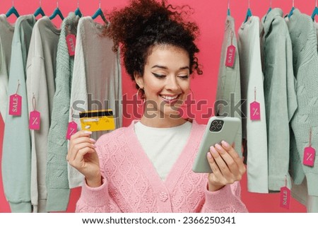 Young woman in sweater stand near clothes rack with tag sale in store showroom using mobile cell phone hold credit bank card shopping online order delivery isolated on plain pink background studio