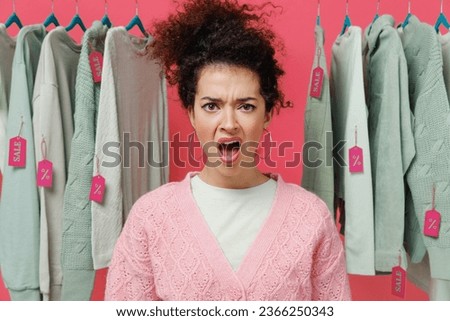 Young sad angry female costumer woman 20s wearing sweater stand near clothes rack with tag sale in store showroom scream look camera with open mouth isolated on plain pink background studio portrait