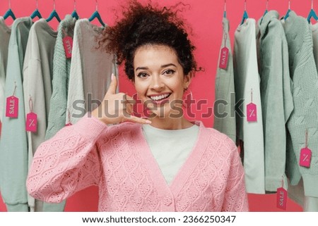 Young fun female costumer woman 20s wear sweater stand near clothes rack with tag sale in store showroom doing phone gesture like says call me back isolated on plain pink background studio portrait
