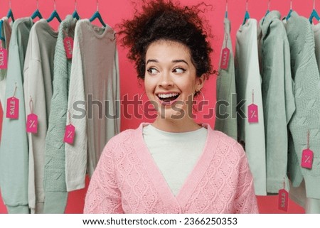 Young smiling happy female costumer woman wear sweater stand near clothes rack with tag sale in store showroom look aside isolated on plain pink background studio portrait. shopping fashion concept