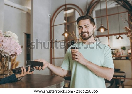 Young man in mint t-shirt buy takeaway paper cup coffee bar counter hold wireless modern bank payment terminal use mobile phone nfc indoors cafe Urban leisure lifestyle catering establishment concept Royalty-Free Stock Photo #2366250365