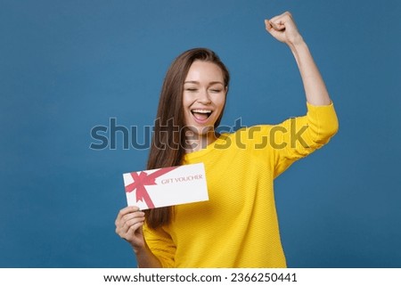 Happy joyful young brunette woman 20s wearing yellow casual clothes posing stand holding in hand gift certificate doing winner gesture keeping eyes closed isolated on blue background studio portrait