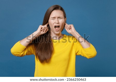 Crazy frustrated displeased young brunette woman 20s wearing yellow casual clothes posing covering ears with fingers keeping eyes closed screaming isolated on blue color background studio portrait