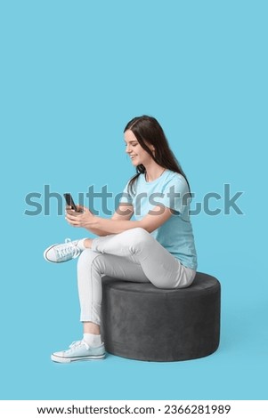 Young woman using mobile phone on pouf against blue background Royalty-Free Stock Photo #2366281989