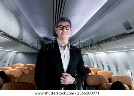 Portrait of happy smiling businessman in black suit, standing on aisle inside airplane, male passenger traveling on business trip by aircraft, businesspeople traveling with airline transportation. Royalty-Free Stock Photo #2366300971