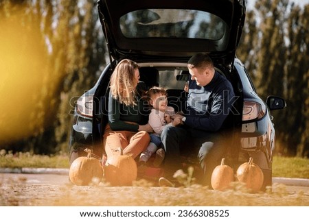 Family day. Happy mom, dad and little child daughter sit at car trunk, look at camera at autumn park with pumpkins, enjoy spend time together at weekend. Parental care and happy carefree childhood.