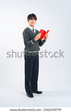 Asian male student standing while reading a book in a white background studio.