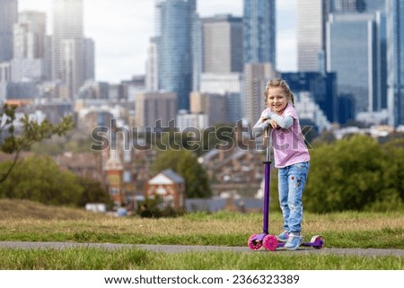 Portrait of a cute, blonde girl with her scooter in a park in front of a urban city skyline