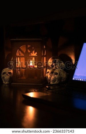 Halloween domestic warm and spooky aesthetic interior decor of vintage lantern with candle and prop skulls near laptop blue lighting display on a table of work space, vertical picture 