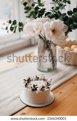 Top view of rustic style white cream cake decorated with fresh flowers, greens and happy birthday inscription on wooden table with natural linen tablecloth. Homemade dessert for birthday party.