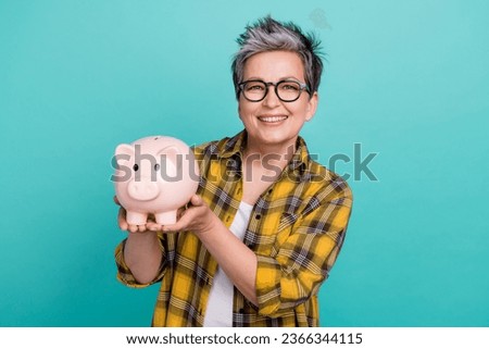 Photo of charming grandmother hold pig bank collect money dressed stylish checkered yellow outfit isolated on aquamarine color background