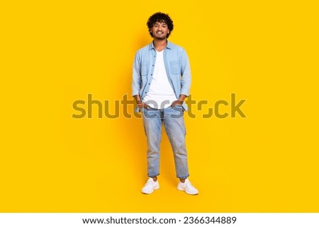 Full size photo of satisfied cool man with curly hairdo dressed denim shirt jeans hold arms in pockets isolated on yellow color background