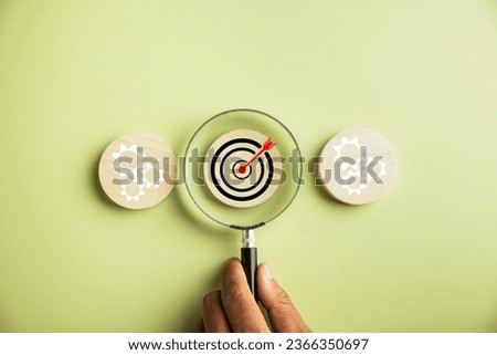 Hand holds a magnifier, focusing on a target icon to analyze objectives and guide management decisions. The image conveys the concept of gear planning, development, and propelling business forward.