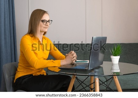 Young focused woman in glasses, an orange blouse, working at a laptop, sitting at a glass table with a notepad for notes. Concept of business, psychology, marketing, training, development.