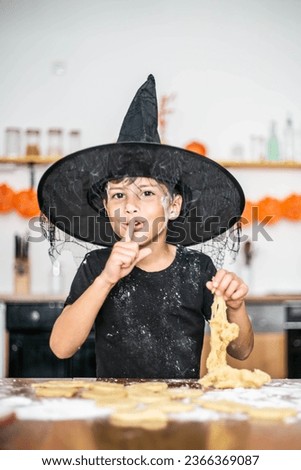Little boy in the kitchen doing a prank. Finger near the mouth as a sign of silence secret, holds the dough while making cookies. Cheerful happy boy in a wizard's hat. Preparing for Halloween.