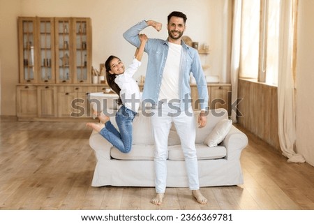 Fun With Dad. Joyful Kid Daughter Hanging On Daddy's Arm While He Showing His Biceps Muscles, Fooling Together At Home Interior, Smiling To Camera. Father's Power And Strength Concept Royalty-Free Stock Photo #2366369781
