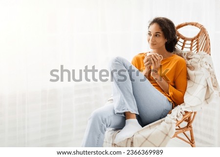 Smiling young woman relaxing in comfortable wicker armchair, holding coffee cup, enjoying cozy weekend morning at home. Middle eastern lady sitting in chair near curtained window, free space