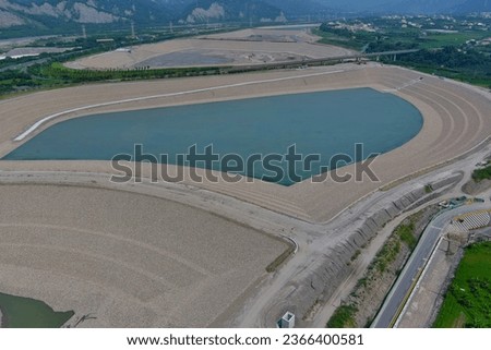 Construction equipment comes into the site. Multi-tiered earthen embankments surround the artificial lake. sediment removal road next to the blue water table, providing access to the lake bottom. Royalty-Free Stock Photo #2366400581