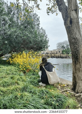 A young woman sits and takes pictures of flowers on the riverbank.