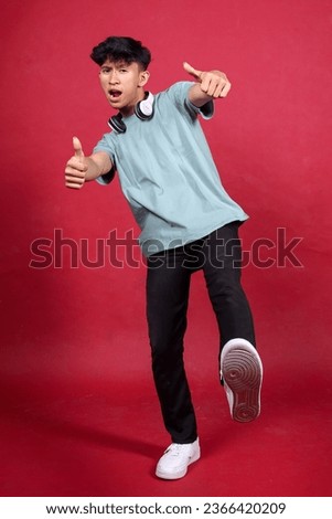 Cool young Asian man with in casual style showing thumbs up to camera and kicking isolated on red crimson background 