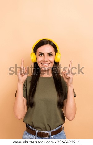 Vertical photo of pretty sweet girl dressed khaki t-shirt enjoying music pointing two fingers up empty space isolated beige color background