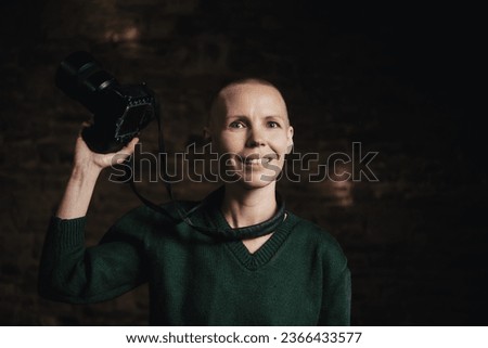 A middle-aged woman photographer holding a camera, smiling, and facing the camera against a dark background