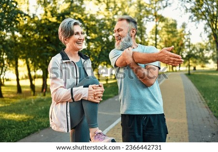 Healthy pensioners exercising outside. Smiling man and woman of senior age standing on paved road and doing physical activities. Slim lady lifting and clutching knee to belly while man stretching arm.